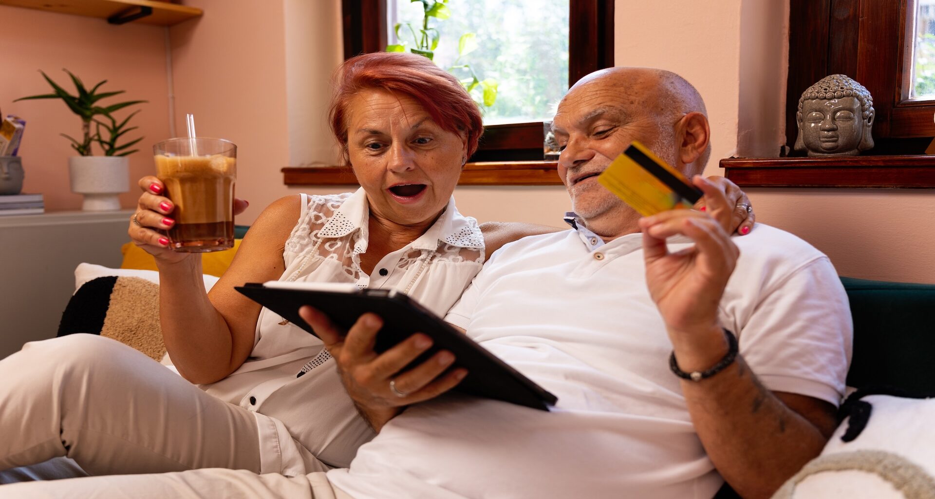A lovely senior couple sits together in their cozy living room, using a tablet for online shopping.