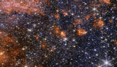 A wide view of a region of space filled with stars and clumps of orange clouds.