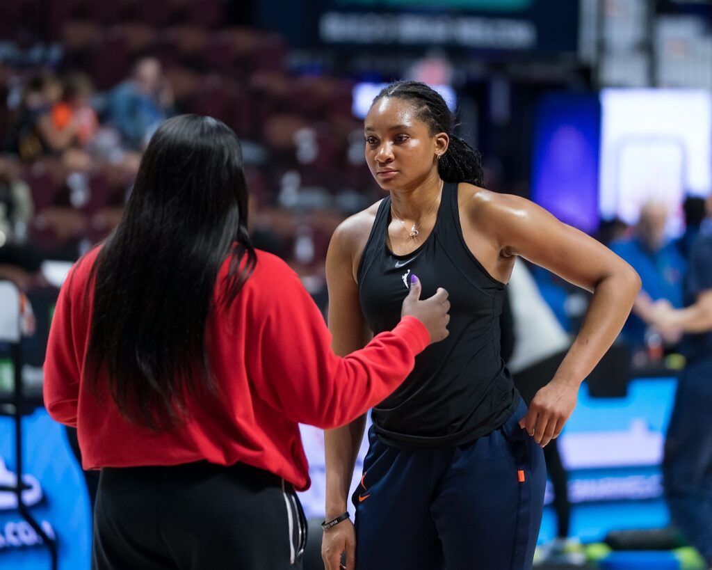 Washington Mystics forward Kiki Iriafen wears a black tank top and navy warm-up pants. She stands with one hand on her hip as she listens to assistant coach Barbara Turner, who's talking to her one-on-one.