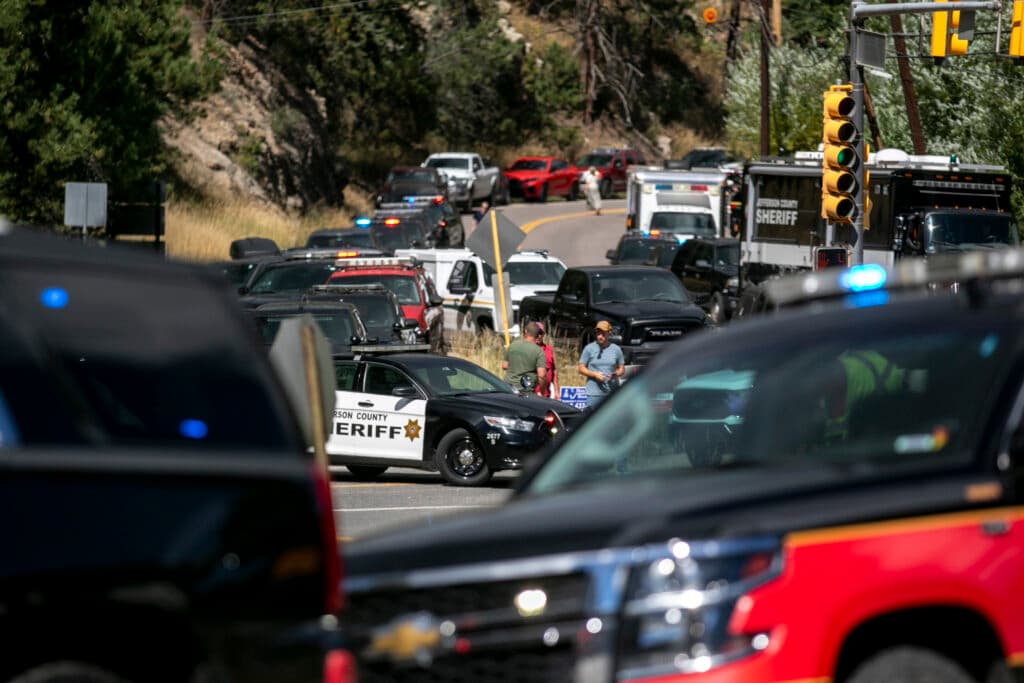 Several police cars and other emergency vehicles block a road lined with trees. Officers and people stand near a county sheriff car.