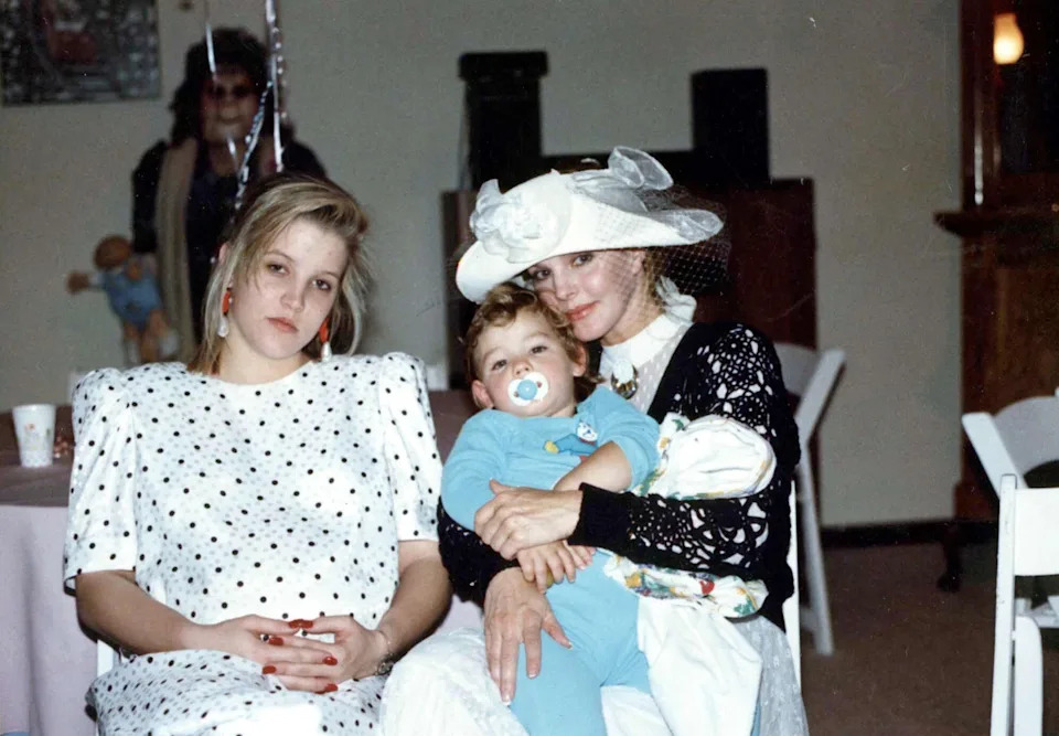 Vintage photo of Lisa Marie Presley sitting beside Priscilla Presley, who holds a baby on her lap while wearing a wide-brimmed hat.