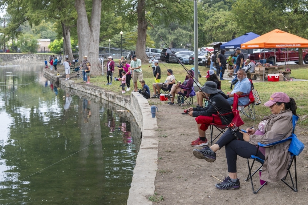 Families fish alongside a river bank with lines dropped into the water waiting for fish to bite.