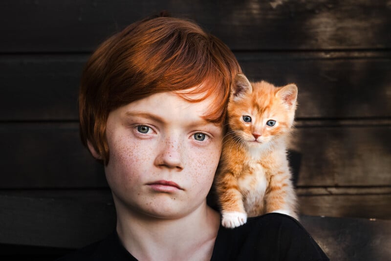 A young child with red hair and freckles looks seriously at the camera, while a small orange kitten with similar coloring sits on their shoulder against a dark wooden background.