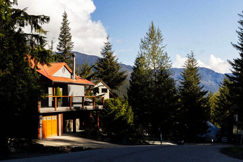 A house with an orange garage door sits among tall pine trees at the edge of a mountain road, with forested slopes and blue sky in the background.
