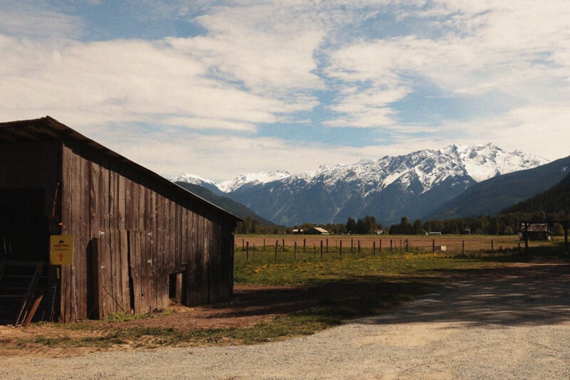 A rustic wooden barn sits beside a gravel road in a rural field, with snow-capped mountains and a partly cloudy sky in the distance.