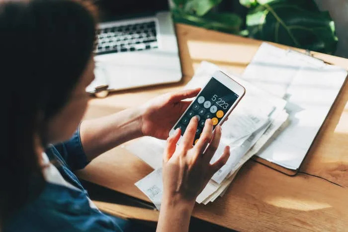 Person using a smartphone calculator, working on finances at a desk with a laptop and paperwork