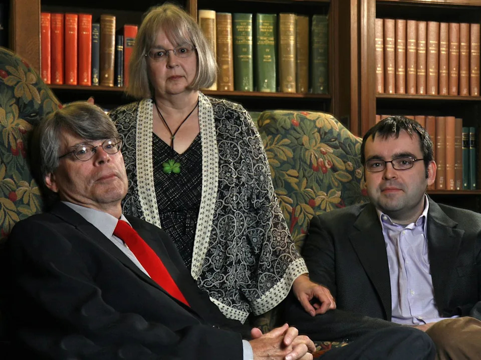 two men sit in high backed chairs as a woman stands in between, all three people look at the camera