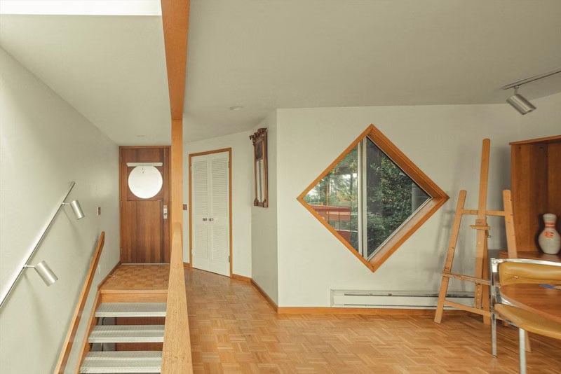 A modern interior hallway features wood flooring, a staircase with a metal handrail, a wooden front door with a round window, a diamond-shaped window, an easel, a small table, and white closet doors.