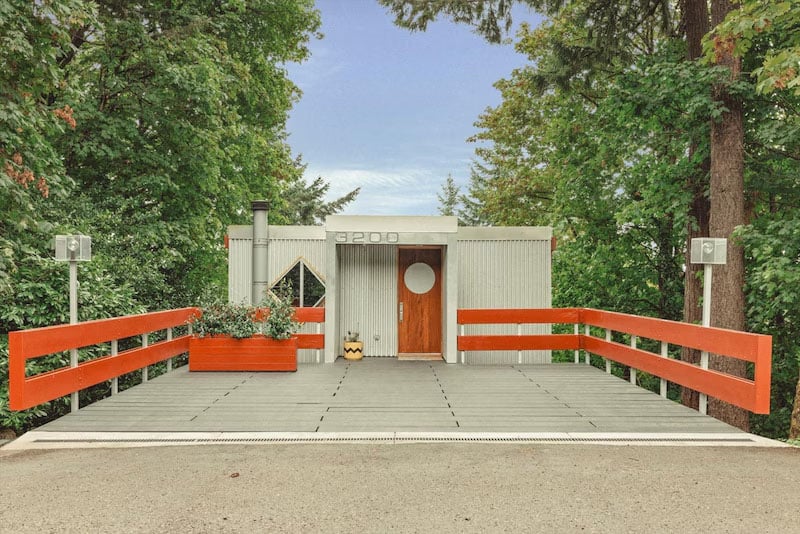 A modern house with a flat roof, gray exterior, and a circular window on the wooden door. It features orange railings and a matching planter, surrounded by green trees. The address "3200" is displayed above the entrance.