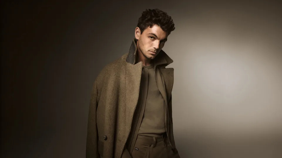 A man in a stylish brown coat and shirt poses against a neutral background, looking confidently at the camera.
