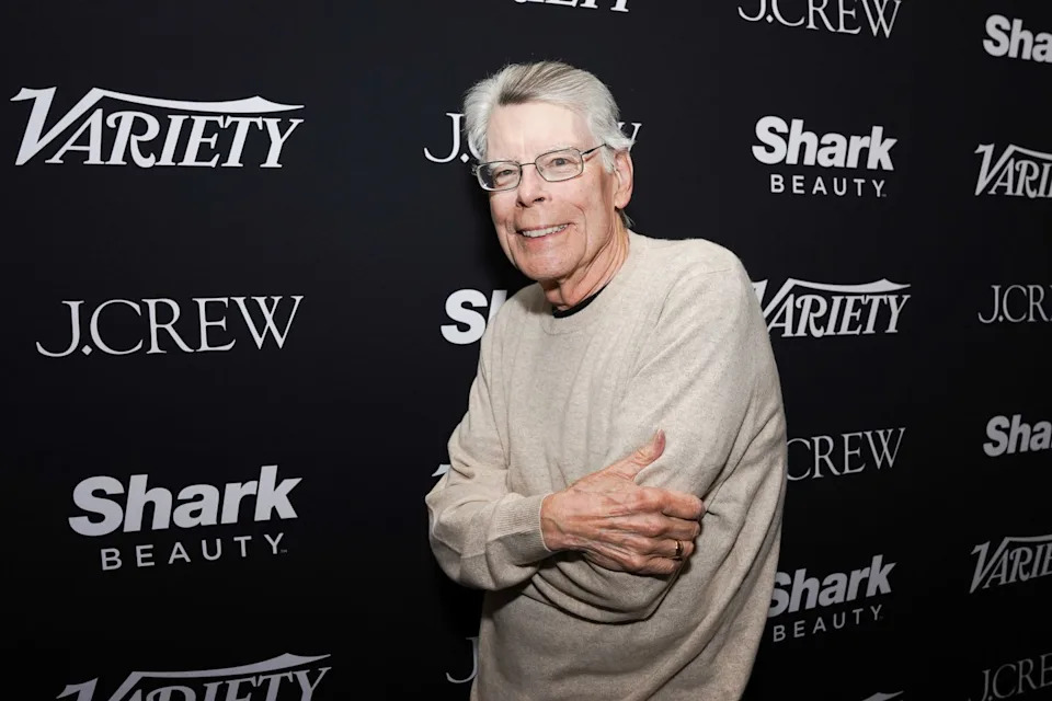 a man in a cream colored long sleeve sweater smiles in front of a black background with logos