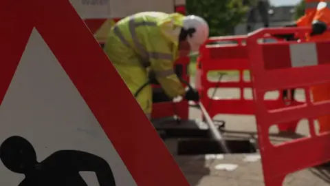 BBC A water company worker in high-viz is clothing is spraying a jet of water from a pressure washer into a sewer. The open manhole is surrounded by red plastic health and safety fencing. 