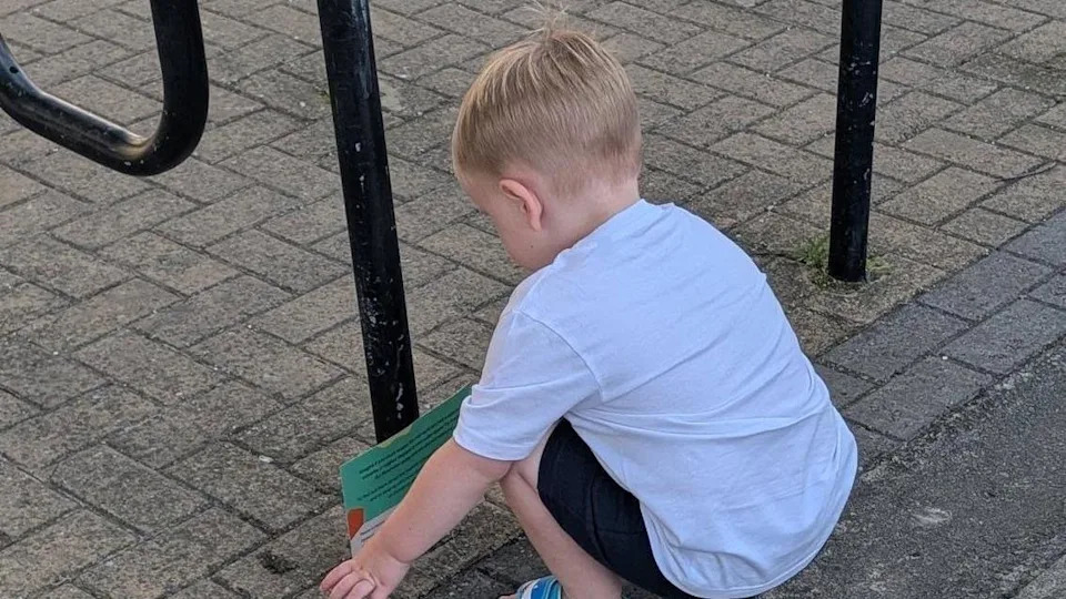 A boy wearing a white T-shirt and black shorts trying to pick up a green voucher placed by the trust by a fence on the ground.