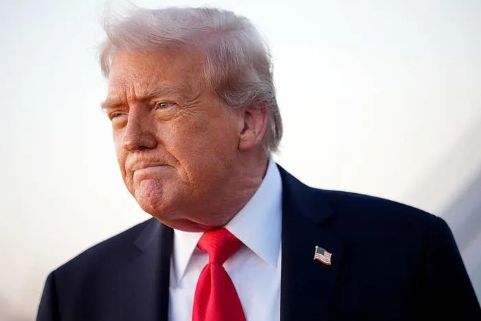 Man in a suit and red tie with an American flag pin looks thoughtfully to the side against a plain background