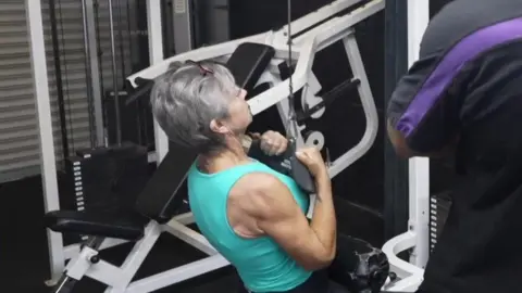 Shaun Whitmore/BBC Ms Smith sits on a bench in front of pulldown machine. She pulls down a weighted handle and holds it in front of her chest. A gym instructor coaches her from the side. 