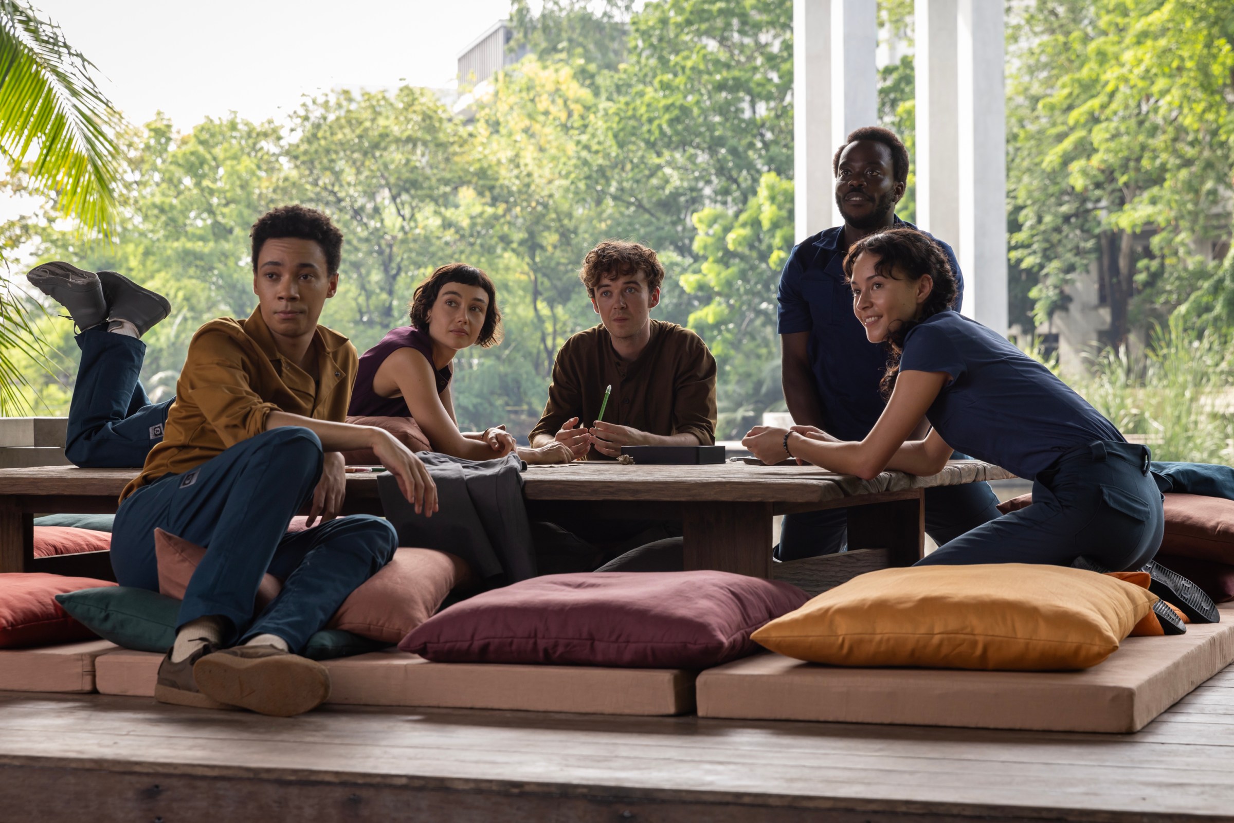 Two women and three men all sitting together at a table that’s close to the ground and surrounded by pillows to sit on. Behind the group is a lush jungle that can be seen through a large glass window.