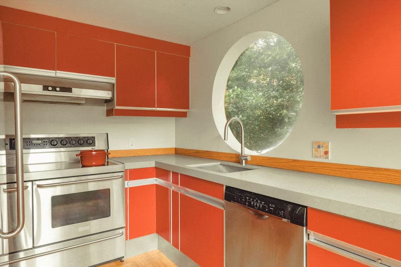 Modern kitchen with red cabinets, stainless steel appliances, a gray countertop, and a round window above the sink that looks out onto greenery. A small red pot sits on the stove.