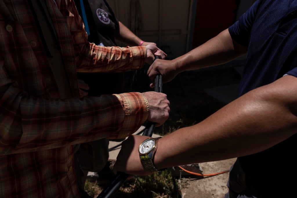 Four people grip a black pipe or rod outdoors, with only their arms and torsos visible in partial sunlight.