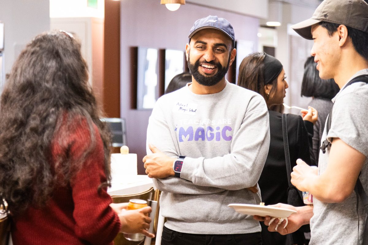 A group of people in casual attire are standing and conversing in a shared indoor space. One person is smiling and wearing a cap, while two others engage with beverages in their hands.