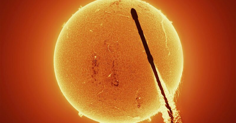 A dark silhouette of a rocket passing in front of the bright, detailed surface of the Sun, with visible solar activity and glowing orange plasma.