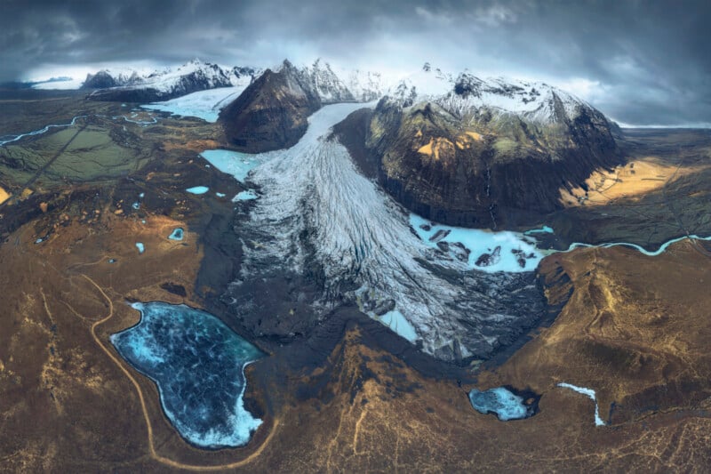 Aerial view of a dramatic glacier flowing between snow-capped mountains, with blue lakes and rivers scattered across a rugged, brown landscape under a cloudy sky.