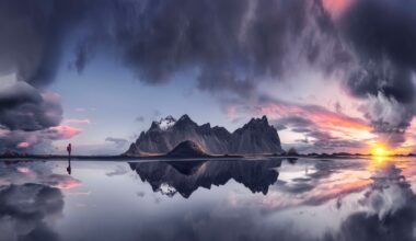 A dramatic landscape with mountains reflected in still water at sunset, clouds fill the sky, and a lone person in red stands to the left, emphasizing the vastness and beauty of the scene.