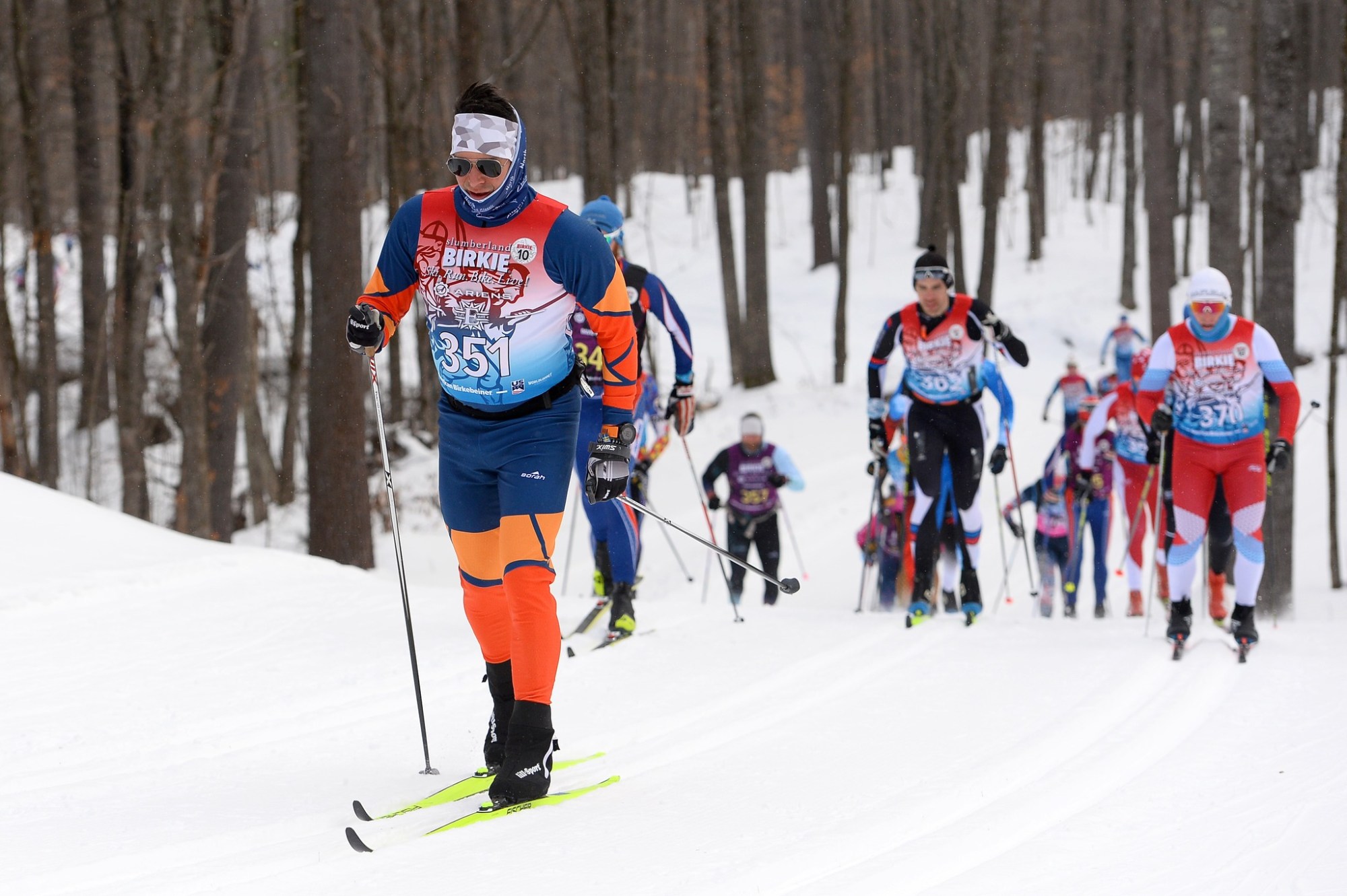 Birkie cross-country skiers ski on snow with trees in background.