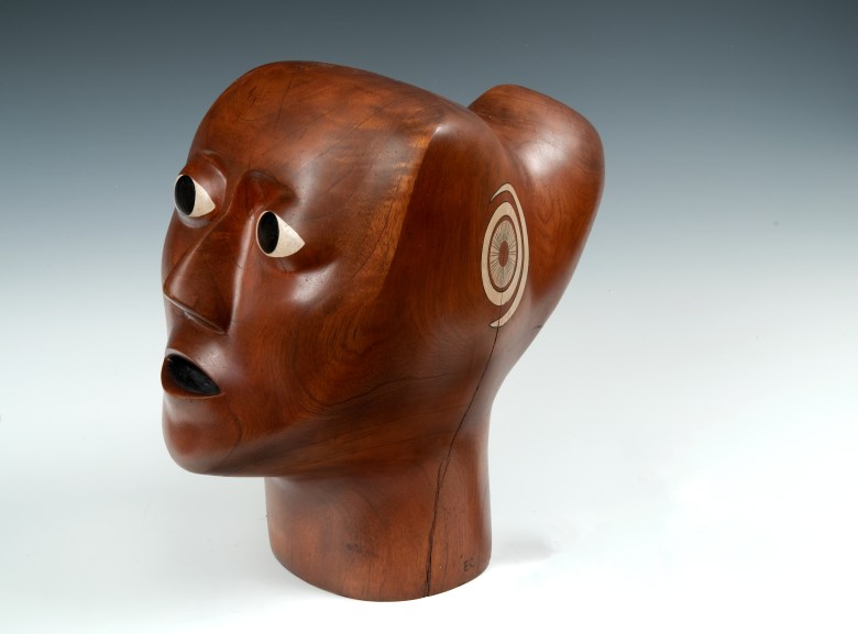 A rich brown wooden bust sculpted in an angular fashion showing the face of a Black woman. Her eyes, lips, and ears are painted, the rest is just the color of the polished wood.