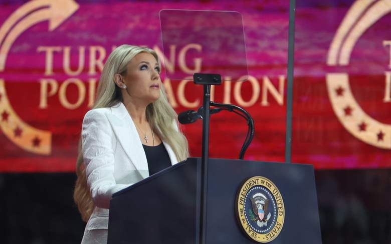A woman with long blond hair speaking at a podium with a presidential seal in front of a red banner.