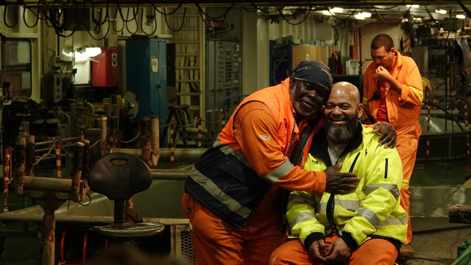 Two workers in safety gear, one wearing an orange jacket and the other in a yellow jacket, share a joyful moment together, smiling and embracing in a workshop setting; another worker in the background is engaged in a task.