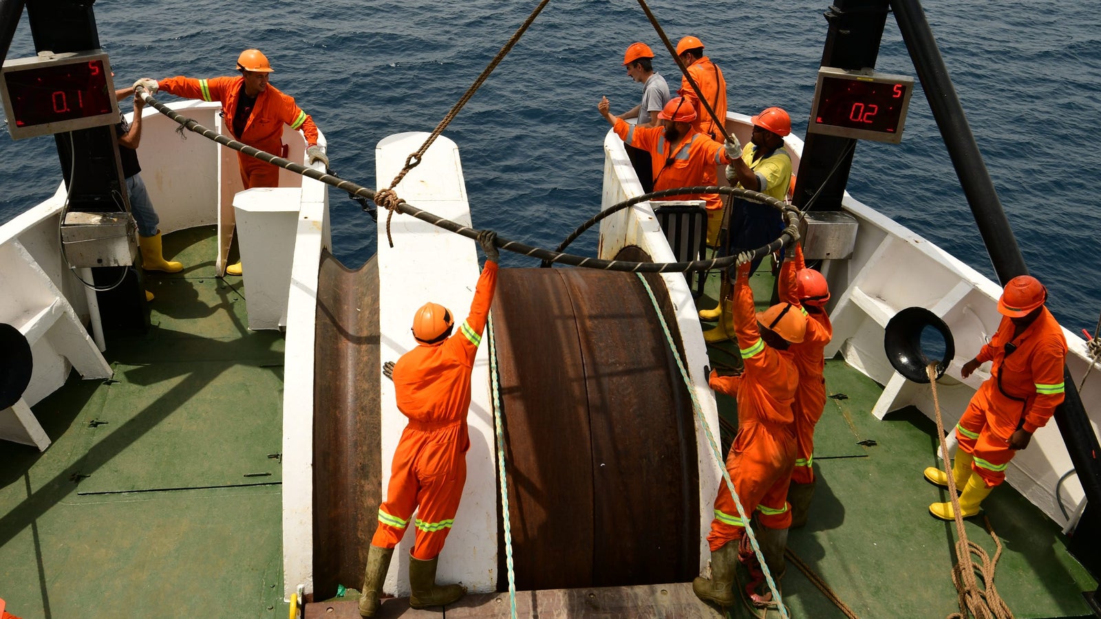A group of seven workers in orange coveralls and hard hats are handling a large, circular metal spool on the deck of a ship, with the ocean visible in the background. Some workers are pulling on ropes while others are preparing to assist, and digital displays are visible showing numerical readings.