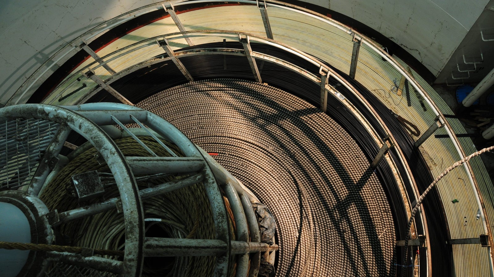 Aerial view of a large circular storage reel with coiled black cables and a separate spool of rope, set against a textured floor, with metal railings visible around the perimeter.