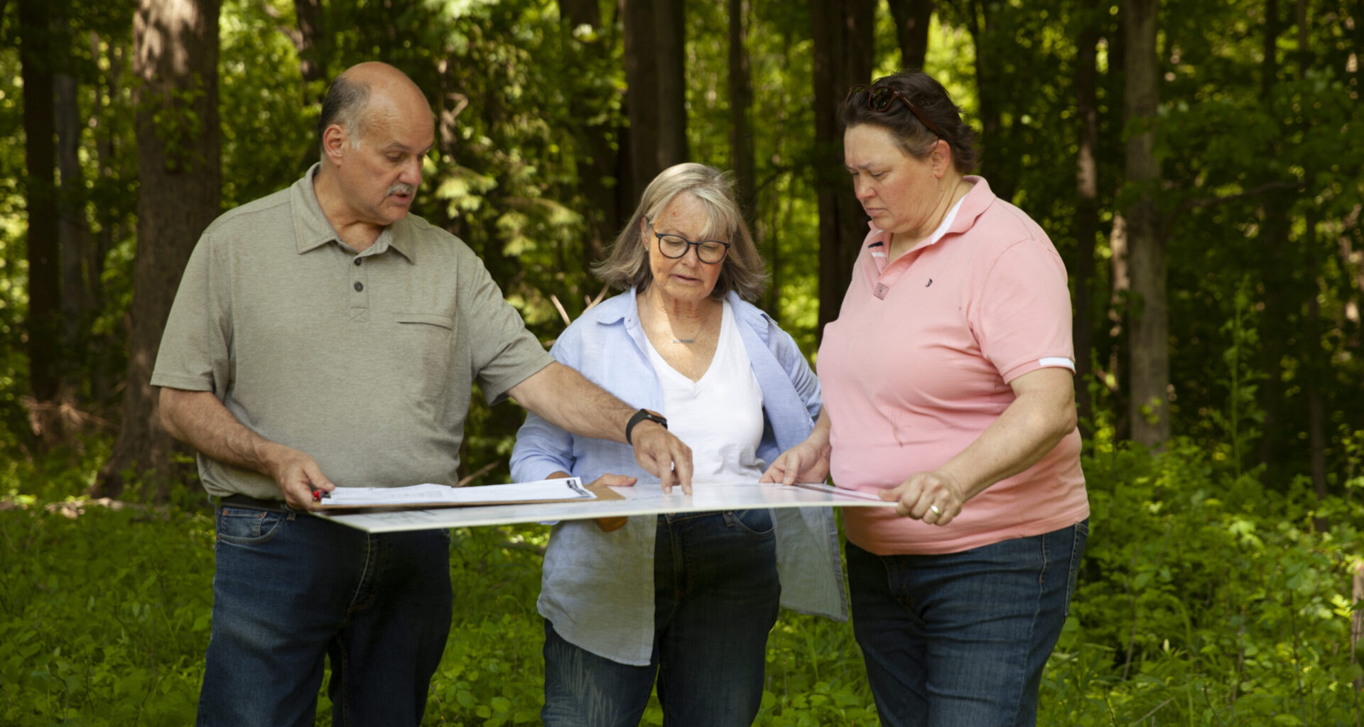 Three adults stand in a wooded area, examining and discussing a large map or blueprint together.