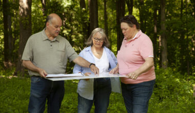 Three adults stand in a wooded area, examining and discussing a large map or blueprint together.