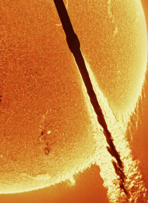 Close-up image of the Sun’s surface, showing swirling textures and bright plasma. A dark, thick line crosses diagonally, casting a shadow over the Sun, possibly from a passing object or stream.