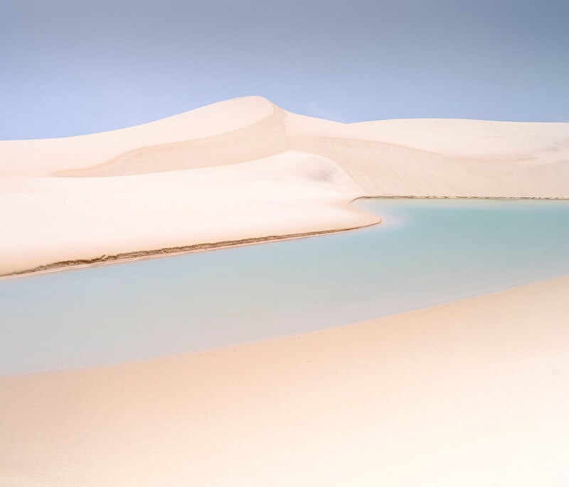 Sweeping white sand dunes border a tranquil, shallow turquoise lagoon under a clear blue sky, creating a serene and minimalistic desert landscape.
