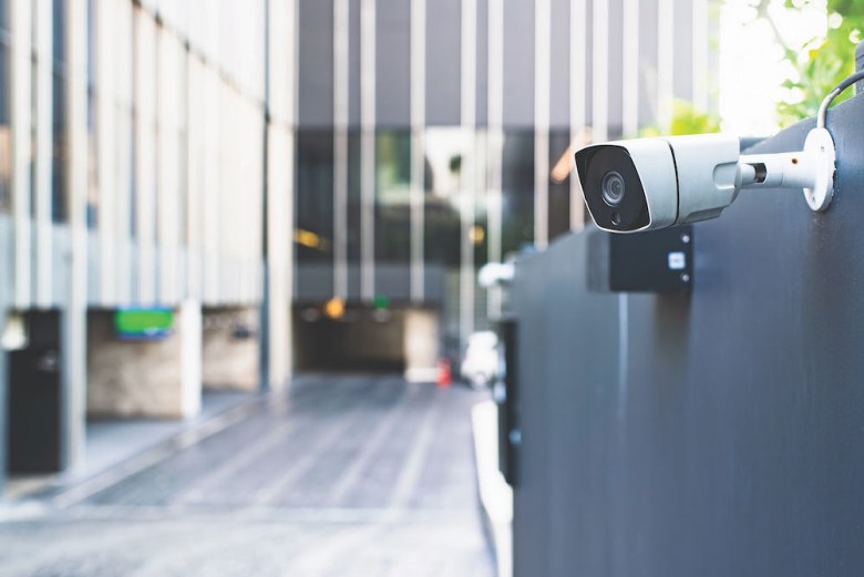 A modern security camera attached to a building's exterior wall oversees an entrance area.