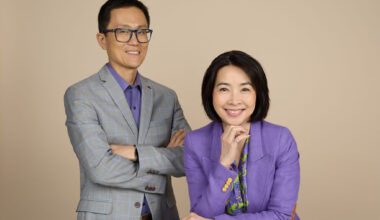 Allen stands beside Eva who is seated at the table. They are smiling at the camera.