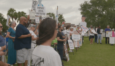 A crowd stands in a park holding signs that say, "Protest Injustice" and "Unmask ICE!"