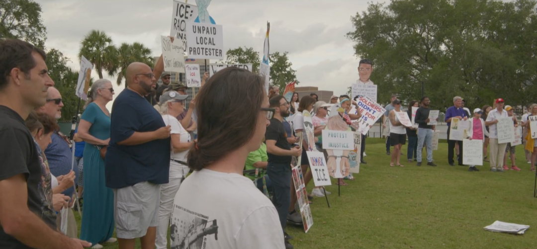 A crowd stands in a park holding signs that say, "Protest Injustice" and "Unmask ICE!"