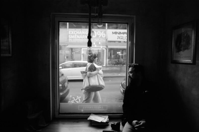 A man sits in a dimly lit room by a window, looking outside. Through the window, a woman carrying bags walks by on the street, and cars and storefront signs are visible in the background.