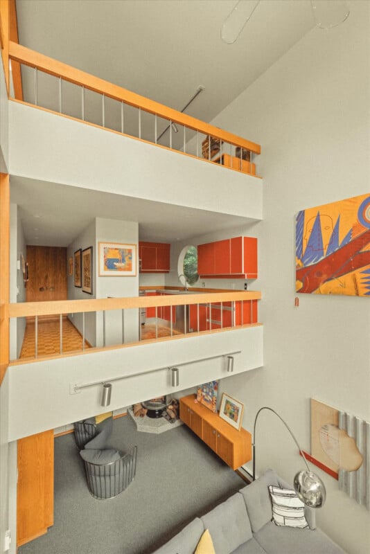A modern, open-concept living space with two loft levels, orange-accented cabinetry, gray furniture, colorful artwork, and large windows, creating a bright and inviting atmosphere.