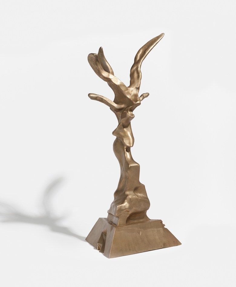 A golden maquette of a metal statue rising from a pyramid-shaped base, into a tall, abstracted, birdlike form.