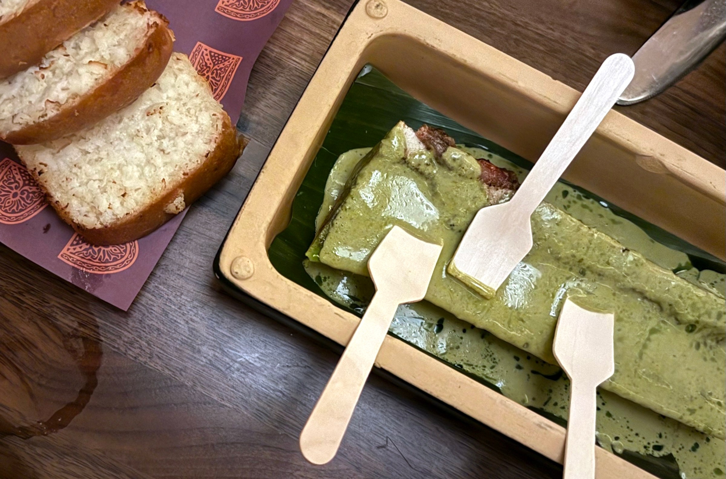 A green-sauced covered bone in a tray with wooden spoons next to slices of bread.