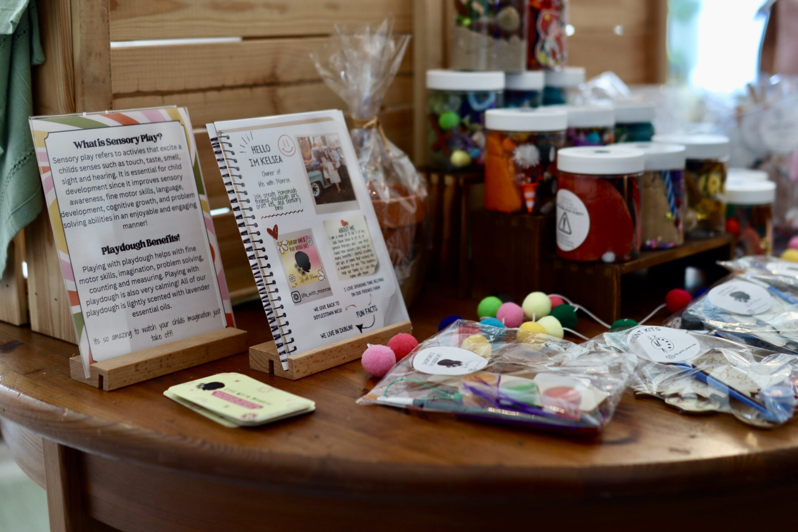 A wooden table displays sensory play kits, jars of craft materials, informative signs, and transparent bags filled with colorful craft supplies.