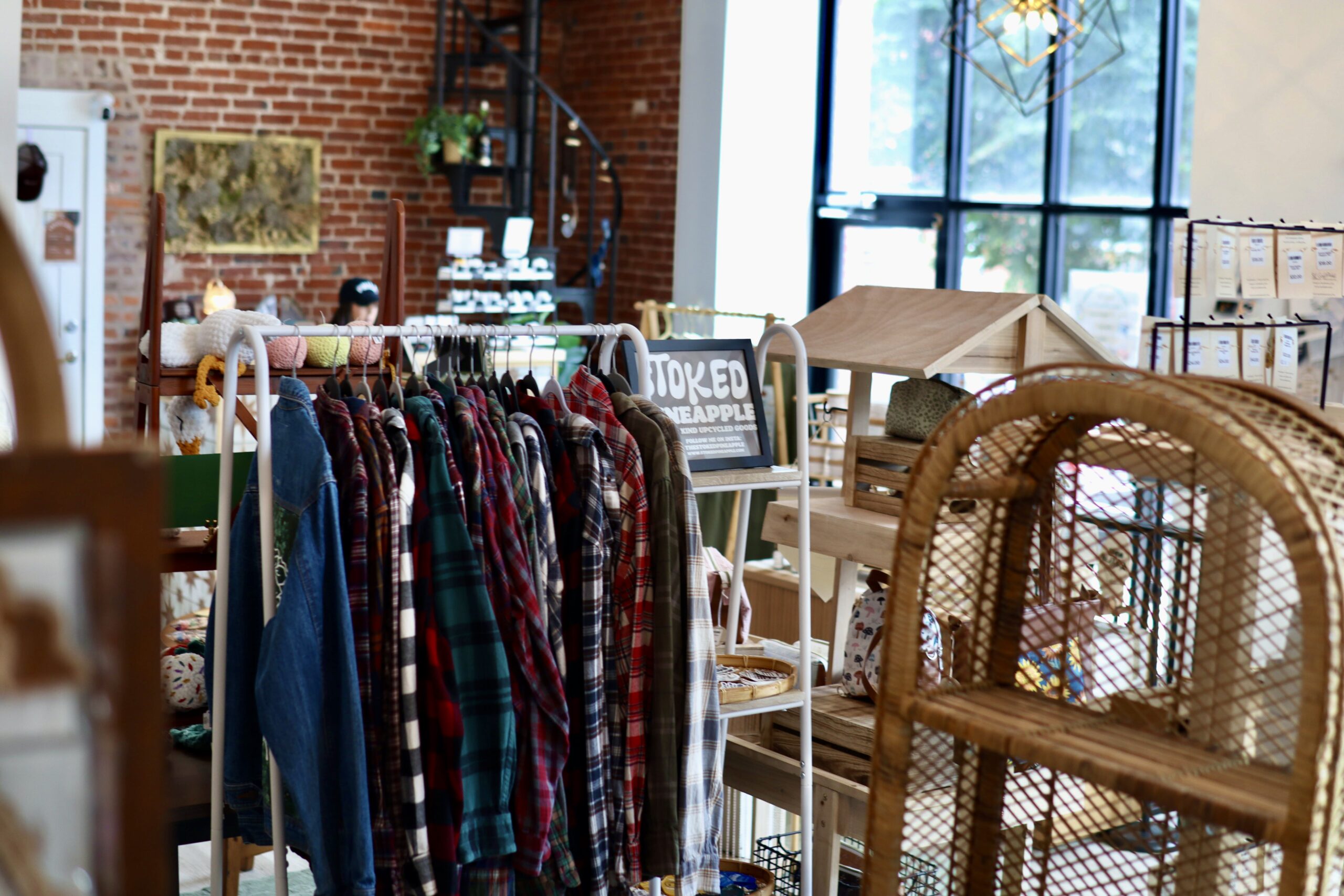 Clothing rack with plaid shirts in a boutique with brick walls, large windows, and various wooden and wicker displays.