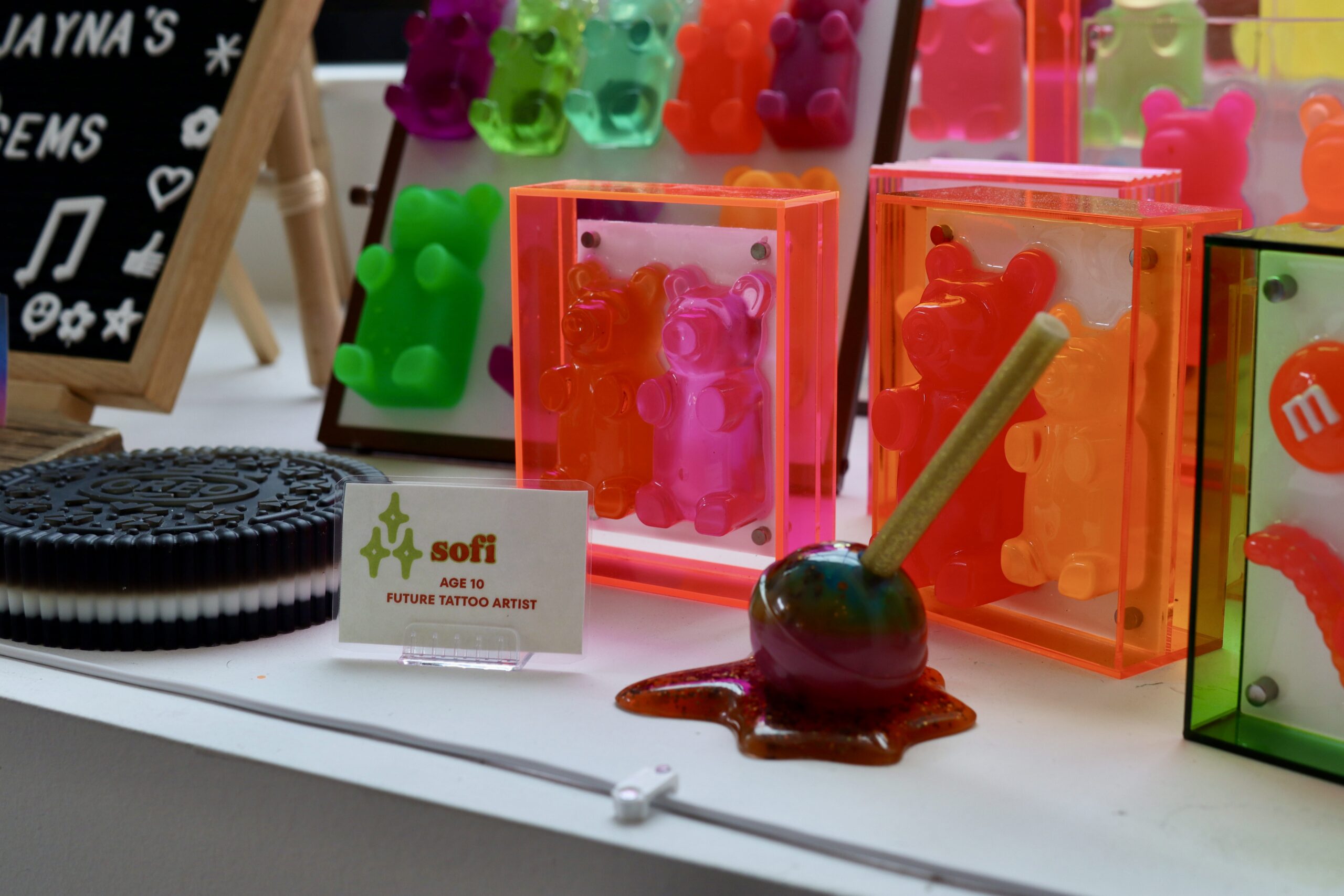 Colorful gummy bear art pieces, a candy apple, an Oreo replica, and a sign reading “sofi age 10 future tattoo artist” displayed on a white surface.
