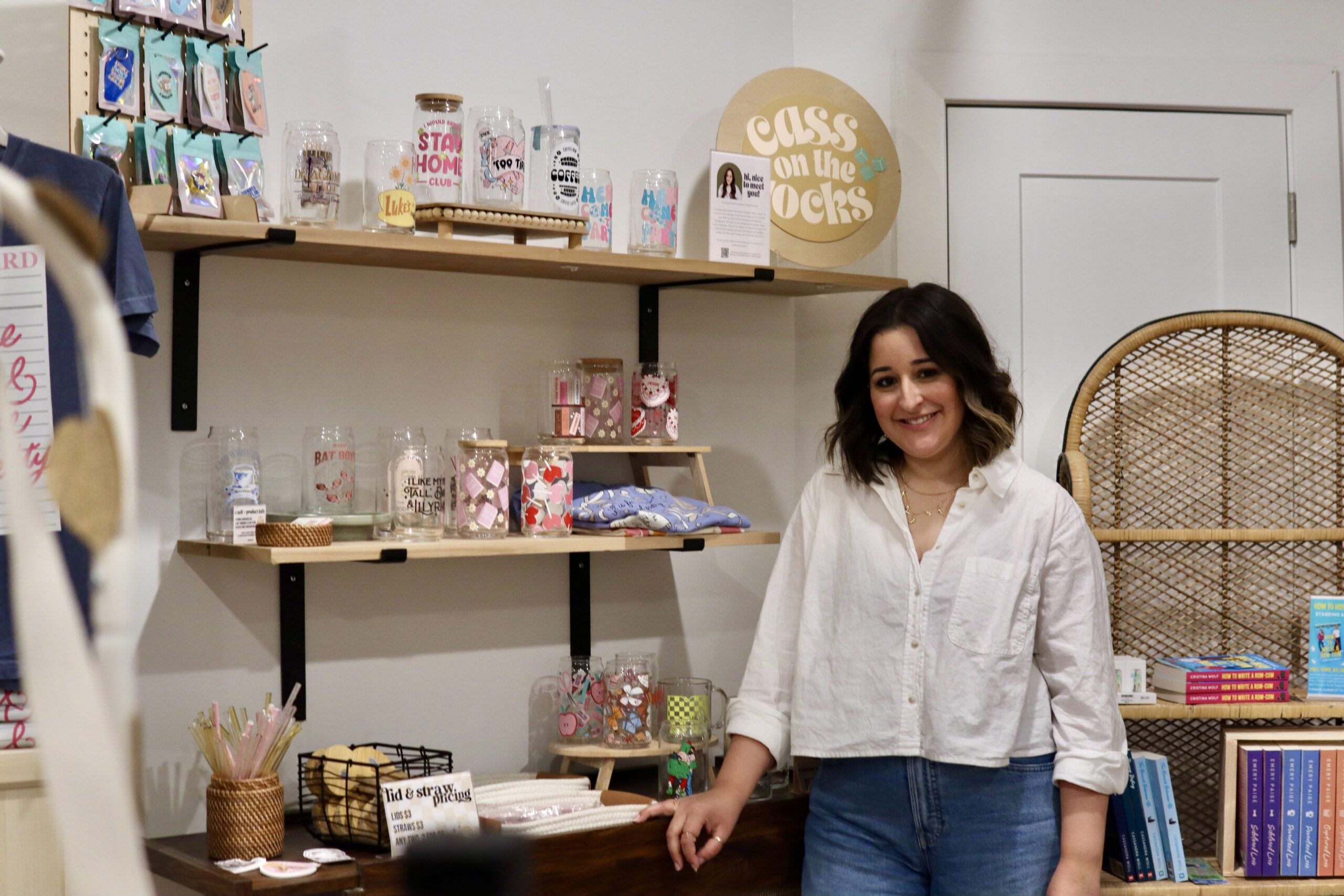 A woman stands in a small shop next to shelves displaying mugs, cups, and gifts. Books are visible on a shelf to the right, and a sign reads "Glass on the Rocks.