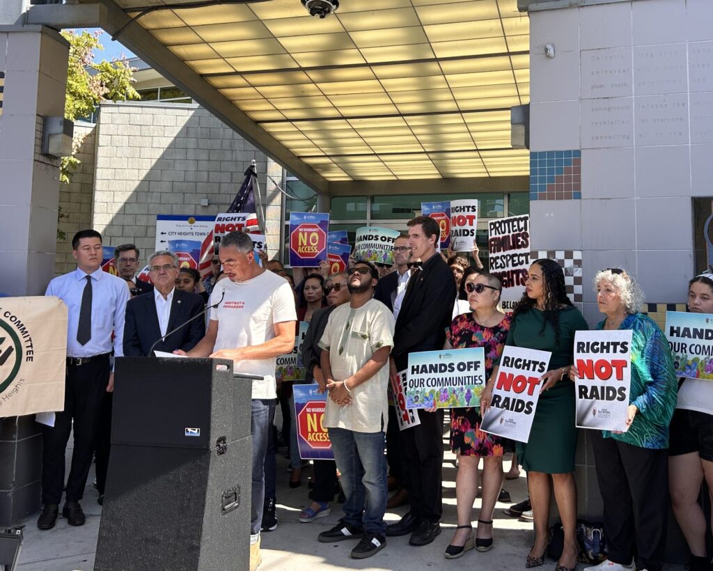 A coalition of politicians at a press conference, with signs that say 'Rights not raids" and "Hands off our communities."
