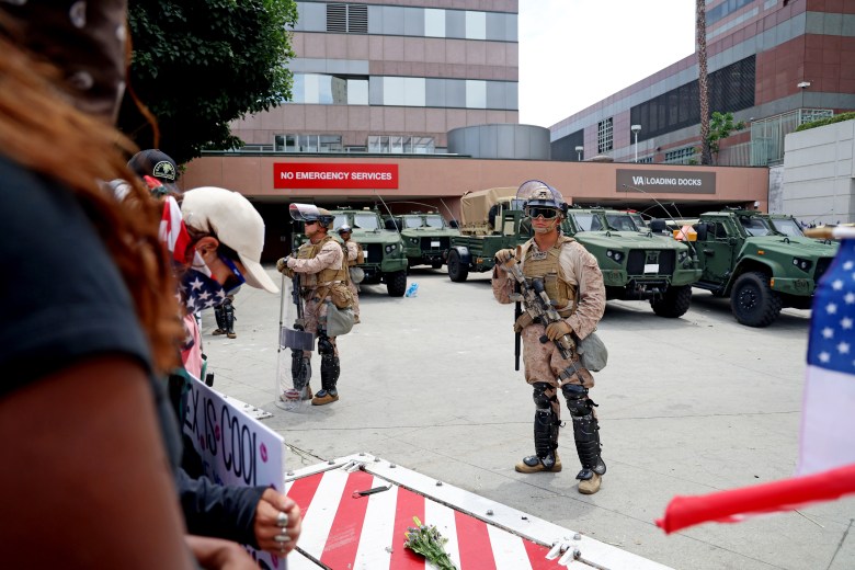 Military personnel in front of armored vehicles, standing near a building with "NO EMERGENCY SERVICES" and "VA LOADING DOCKS" signs.